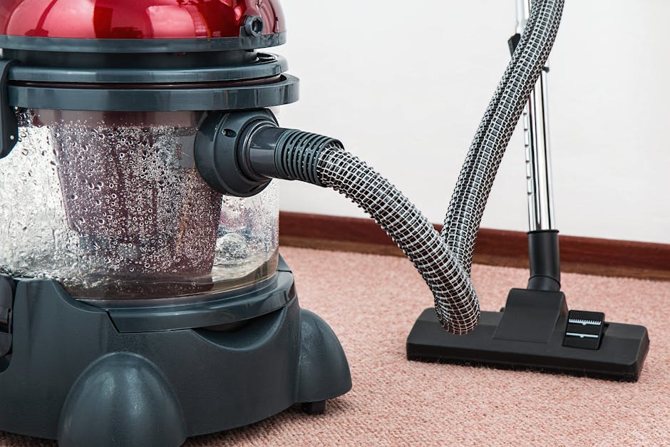 A person operating a yellow portable carpet cleaning machine with a black grill, positioned on a multicolored, patterned carpet in a room with warm wooden floorboards visible in the background. The ro
