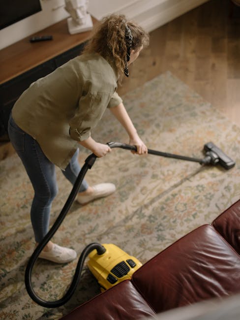 Close-up image of a person using a yellow and black vacuum cleaner on a patterned carpet in a residential setting. The person's hands are holding a white cleaning cloth, inserting it into the vacuum's