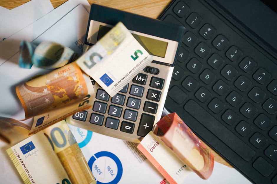 A close-up image showing a calculator with a Euro banknote draped over it, surrounded by scattered Euro banknotes of various denominations on a wooden desk. To the right, there is a black computer key